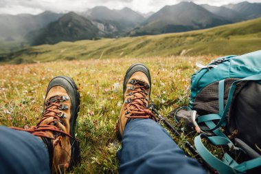 Yükseklerdeki dağların tepesinde sırt çantasıyla gezen bir kadın, çimlerin üzerinde oturuyor.