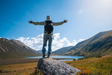 Sırt çantalı bir kadın yüksek irtifada dağ tepesinde yürüyüş yapıyor. Uzakta bir göl var.