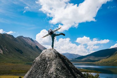 Sırt çantalı bir kadın yüksek irtifada dağ tepesinde yürüyüş yapıyor. Uzakta bir göl var. 