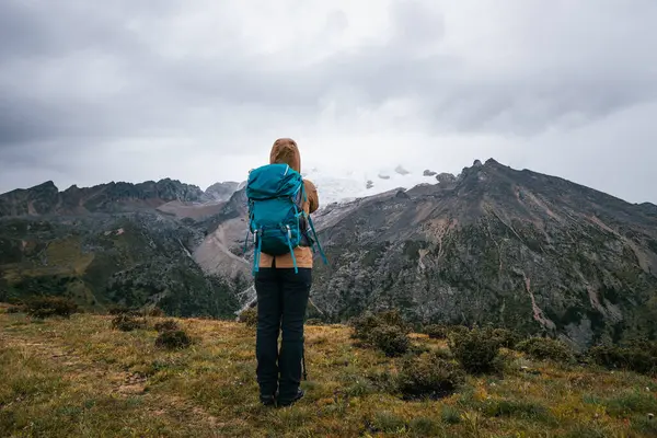 Sırt çantalı bir kadın yüksek irtifada dağ yürüyüşü yapıyor. Yağmurda karla kaplı dağa bakıyor. 