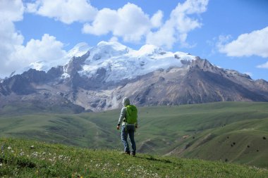 Sırt çantalı bir kadın otlakta ve karlı dağlarda yürüyüş yapıyor.