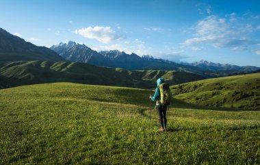 Sırt çantalı bir kadın çayırlarda yürüyüş yapıyor. Dağların tepesinde kar manzarasının tadını çıkarıyor.