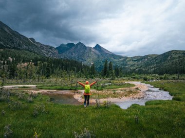 Sırt çantalı bir kadın otlakta ve orman dağlarında yürüyüş yapıyor.  