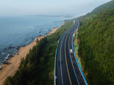 Bir sahil otoyolu, kumlu bir sahil ve diğer tarafında yeşil bir yamaç bulunan sakin bir okyanus boyunca kıvrılır..