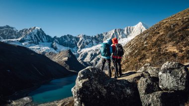 Sırt çantalı iki yürüyüşçü bir kayanın üzerinde durmuş açık mavi gökyüzünün altındaki karlı dağlara bakıyor..
