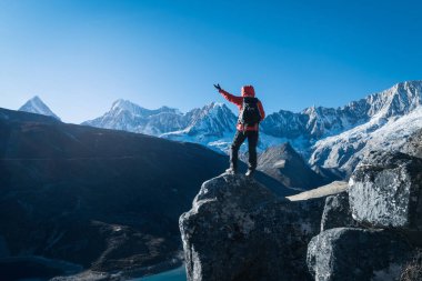 Yürüyüşçü, turkuaz bir göle ve karlı tepelere bakan kayalık bir dağ sırtında duruyor.