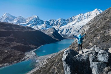Ceketli yalnız yürüyüşçü, turkuaz bir göle ve karlı tepelere bakan kayalık bir dağ sırtında duruyor.