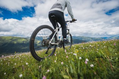 Uçsuz bucaksız gökyüzünün altındaki engebeli dağlara bakan yeşil bir yamaçta bisiklet pedalları.