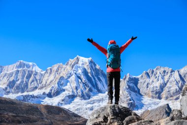 Yürüyüşçü, turkuaz bir göle ve karlı tepelere bakan kayalık bir dağ sırtında duruyor.