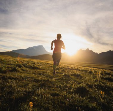Kadın gün batımında yüksek irtifada çayırlarda koşuyor. 