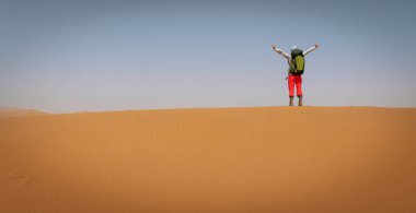 Sırt çantalı bir kadın çölde yürüyor, ellerini havaya kaldırıyor, özgürlüğü selamlıyor.