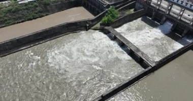 Bir barajın beton sızıntılarından akan çalkantılı beyaz suyun havadan aşağı görüntüsü.