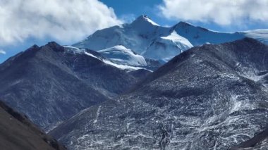 Karla kaplı dağlar ve beyaz bulutlu mavi gökyüzünün altındaki karanlık yamaçlar..