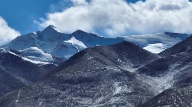 Karla kaplı dağlar ve beyaz bulutlu mavi gökyüzünün altındaki karanlık yamaçlar..