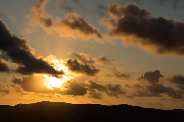Dağ siluetlerinin günbatımı. Bulutlar, alacakaranlıkta güneşin altın ışıklarıyla aydınlanıyor. Fuerteventura, Kanarya Adaları, İspanya