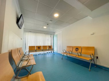 Bekleme odası. Oturmak için ahşap banklar. Mavi katlı bir oda. Duvardaki düz ekran televizyon. Arka planda perdeleri olan pencereler. Işıklı beyaz tavan. Tenerife, Kanarya Adaları, İspanya