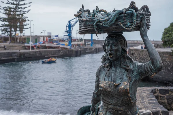 Metal statue of an ancient fisherwoman, with her mouth open, arm raised holding a basket over her head, from which octopus oars are coming out. Pier in the background, with ocean, crane and boats. Puerto de la Cruz, Tenerife, Canary Islands, Spain.