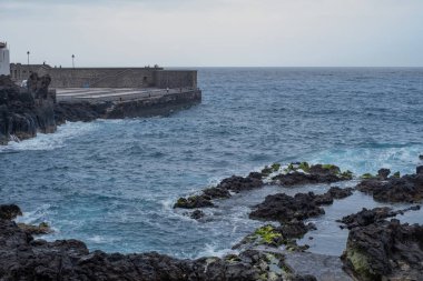 Dibinde bir iskelesi olan doğal havuzlar oluşturan kayalar. Dalgalar uçurumun volkanik kayalarına çarpıyor. Dalgalı okyanus. Siyah kayalar. Puerto de la Cruz, Tenerife, Kanarya Adaları, İspanya.