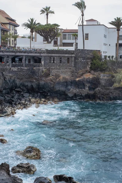 stock image View of the houses on a volcanic rock cliff. Waves breaking against the volcanic rocks of the cliff. Rough ocean. Black rocks. Puerto de la Cruz, Tenerife, Canary Islands, Spain.