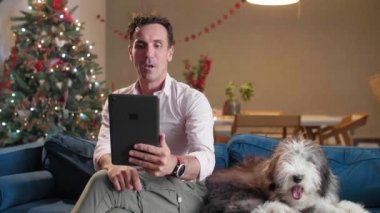 Adam oturma odasındaki kanepede oturuyor Noel süsleriyle, köpeğini okşuyor ve tablet üzerinde çalışıyor..