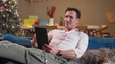 Adam oturma odasındaki kanepede oturuyor Noel süsleriyle, köpeğini okşuyor ve tablet üzerinde çalışıyor..