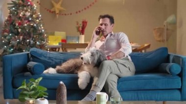 Adam telefonda konuşurken kanepede oturuyor. Aynı zamanda, o da kanepede yatan köpeğini okşar. Oturma odası Noel ruhuyla süslenmiş..