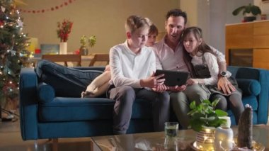 Bir baba, çocuklarının oturma odasında kanepede otururken onlar tabletten bir şey izliyorlar. Oda şenlikli bir ruhla dekore edildi. Gelecek Noel 'le birlikte..