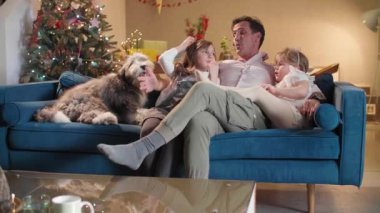 Bir baba oturma odasındaki kanepede iki kızı ve köpeğiyle oturuyor. Evlerinde uzanırken gülümsüyorlar ve neşeliler. Oda Noel ruhuyla süslenmiş..