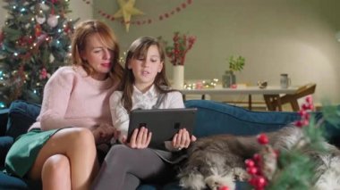 Anne kızı ve bir köpekle kanepede oturuyor. Oda Noel ruhu ile süslenmiştir. Tablette bir şey izlerken gülümsüyorlar ve neşeliler..