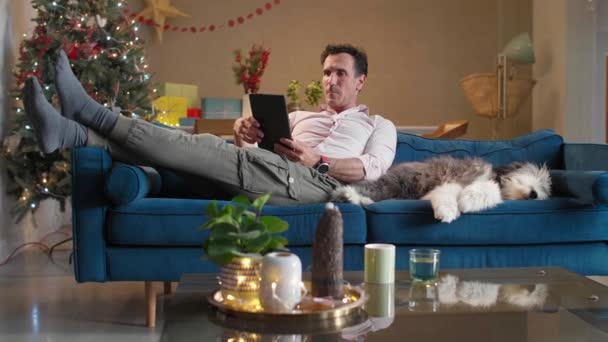 Чоловік Сидить Дивані Вітальні Різдвяними Прикрасами Гладить Собаку Працює Планшеті — стокове відео