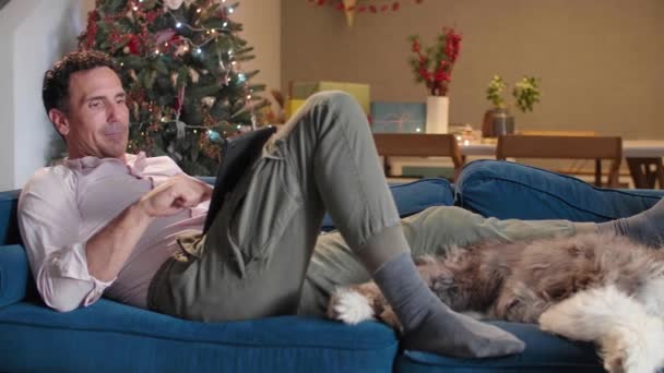 男人们坐在客厅的沙发上 摆放着圣诞装饰品 爱抚着他的狗 还在做平板电脑 — 图库视频影像