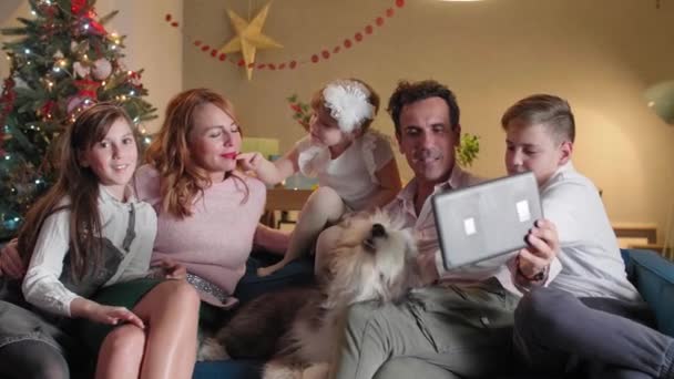 幸せな家族はリビングルームのソファに座っており タブレット経由でビデオ通話をしています 部屋はクリスマスの精神で飾られています かれらの犬はソファの上にいて — ストック動画