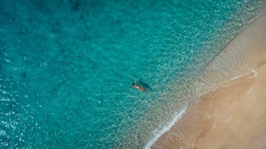 Yüzeysel deniz suyunda uzanmış, kumlu kumsalın ve yumuşak turkuaz okyanus dalgasının tadını çıkaran mayolu güzel mutlu kadının hava manzarası. Yaz mevsiminde Lefkada adasındaki Egremni sahilinde tropikal deniz..