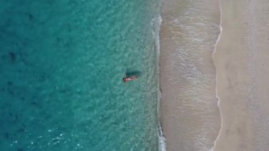 Aerial view of beautiful happy woman in swimsuit laying in the shallow sea water, enjoying sandy beach and soft turquoise ocean wave. Tropical sea in summer season on Egremni beach on Lefkada island.