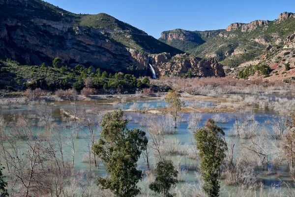 Beautiful landscape of the waterfall near the old town of Domeo on a rocky mountain full of vegetation and the Turia river with three trees in the foreground, in Valencia, Spain
