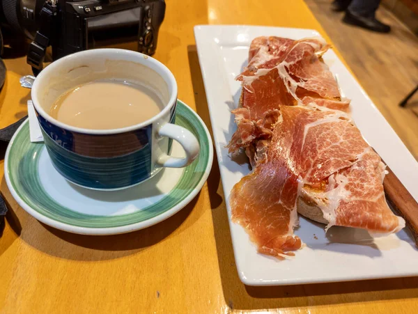 Beautiful image of the best Spanish breakfast, a toast of soft and crunchy homemade bread with grated tomato and a good serrano ham on top accompanied with a coffee with milk, in the background you can see a camera