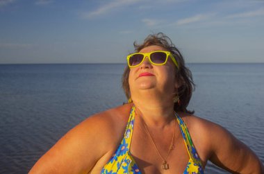 Mavi mayo giymiş orta yaşlı bir kadın kolları açık ve güneşlenerek kıyıda duruyor.