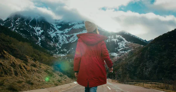 stock image Woman in red jacket on winter travel adventure walk on empty mountain road surrounded by beautiful nature. Tourist female on hiking trip explores earth