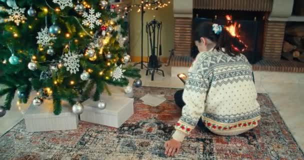 若い大人の女性は自宅で携帯電話を使用してサイドクリスマスツリーで暖炉の前に座っている — ストック動画