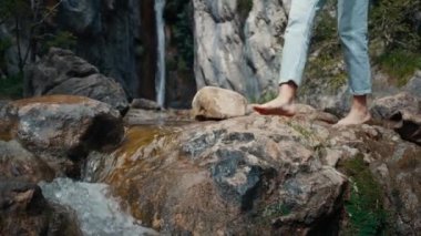 Çıplak ayaklı kadın turist, arka planda şelaleyle dağ nehrinden geçen kayalıklarda yürüyor. Sağlıklı yaşam tarzı kavramı ve doğa üzerine seyahat macerası. Tatilde ayak izlerinin dinlenmesi