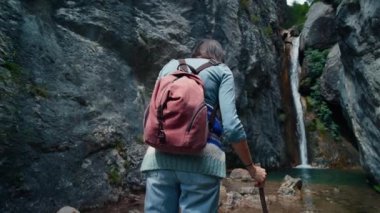 Dişi gezgin sırt çantasıyla dağ yolunda kayalıklarda yürüyor. Sırt çantalı turist kadın dağ nehri şelalesine yaklaşıyor. 