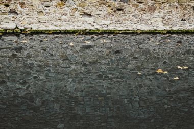 Nehrin suyunda eş merkezli dalga deseni ve nehir boyunca uzanan antik taş duvarın yansıması. Suyun yüzeyinde küçük yapraklar yüzer..