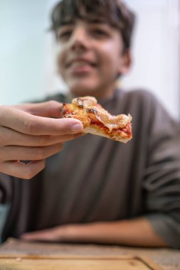 Genç bir çocuğun bir dilim pizzayı tutarken yakın plan fotoğrafı. Gülümseyen çocuğun görüntüsü nazikçe bulanıklaşıyor..