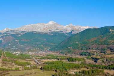 Göz kamaştırıcı bir dağ silsilesi kar kaplı tepeleri parlak mavi gökyüzüne karşı sergiliyor. Aşağıda, yeşil vadiler ve ağaçlar tuhaf bir köyün yakınındaki sonbaharda doğanın güzelliğini yansıtıyor..