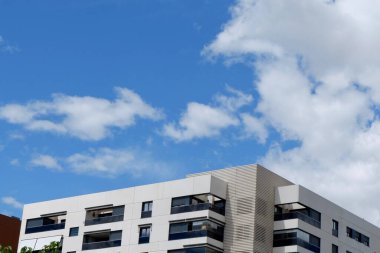 Parlak ve modern apartman binası dağınık kabarık bulutlarla dolu canlı mavi bir gökyüzüne karşı duruyor. Mimaride, güneş ışığını yansıtan temiz çizgiler ve geniş pencereler yer alıyor..