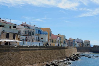 Parlak güneşli bir günde kıyı şeridini kaplayan pastel renkli binalar ön plana çıkıyor. Dalgalar kayalık kıyılara çarpar ve deniz kenarı kasabasında sakin bir atmosfer yaratır..