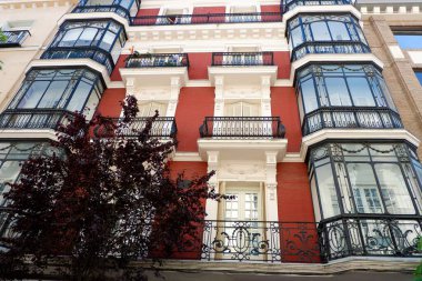 Renkli kırmızı dış görünüş, sayısız cam balkon içeren kentsel bir apartmanı süslüyor, berrak gökyüzü altında karmaşık mimari detaylar sergiliyor..