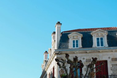 Parlak güneş ışığı, karmaşık taş işçiliğini ve iyi korunmuş mimarisini sergileyerek, tarihi bir binanın zarif yüzünü aydınlatıyor. Çatıda klasik yatakhane pencereleri ve şık bir kaplama var. Altında, budanmış ağaçlar 