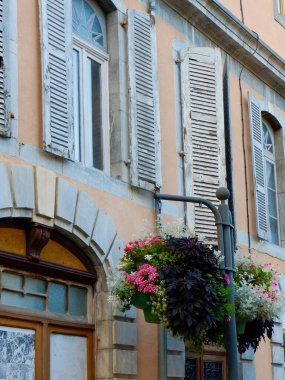 Bir binada ahşap panjurlarla süslenmiş soluk duvarlar ve elektrik direğinden sarkan canlı çiçek sepetleri yer alıyor. Sahne hayat dolu, ılık güneş ışığında çiçek sergileniyor..