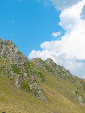 Kayalık dağ arazisi yemyeşil ve dağınık bulutlu açık mavi bir gökyüzüne sahiptir. Açık hava aktiviteleri ve keşifleri için ideal sakin bir doğal ortam..
